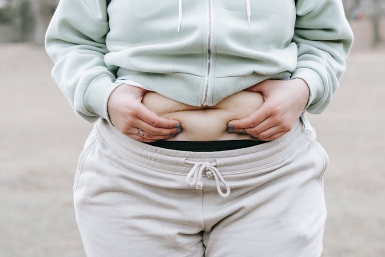 Crop anonymous plus sized female in casual hoodie touching and showing fat on belly while standing in park