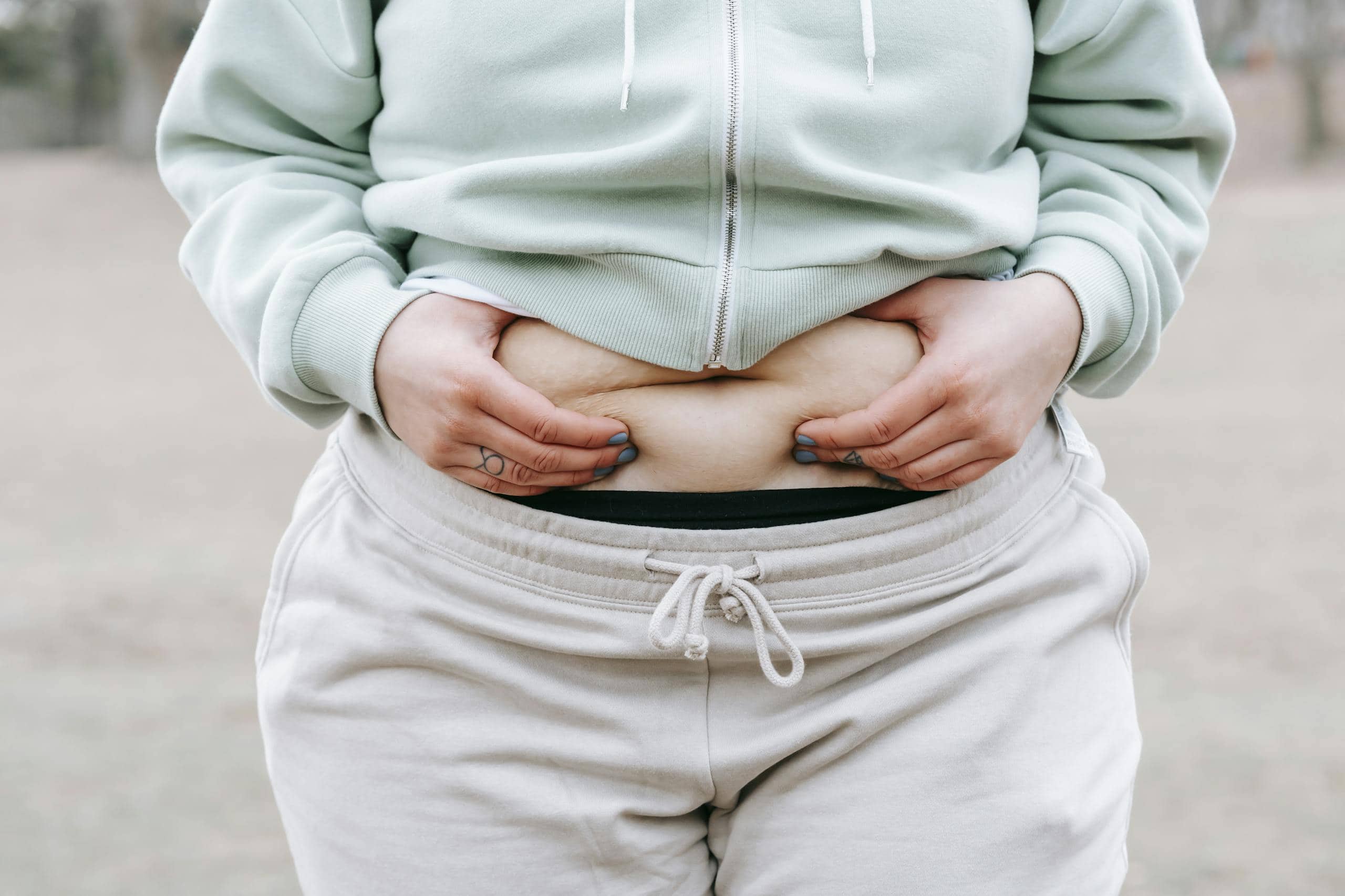 Crop anonymous plus sized female in casual hoodie touching and showing fat on belly while standing in park