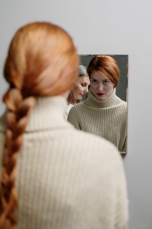 A woman with auburn braided hair gazes into a mirror reflecting her and an elder woman.