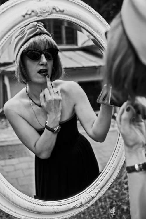 Black and white of gorgeous young slender female in black off shoulder dress and sunglasses demonstrating Fuck You hand gesture while looking at oval mirror