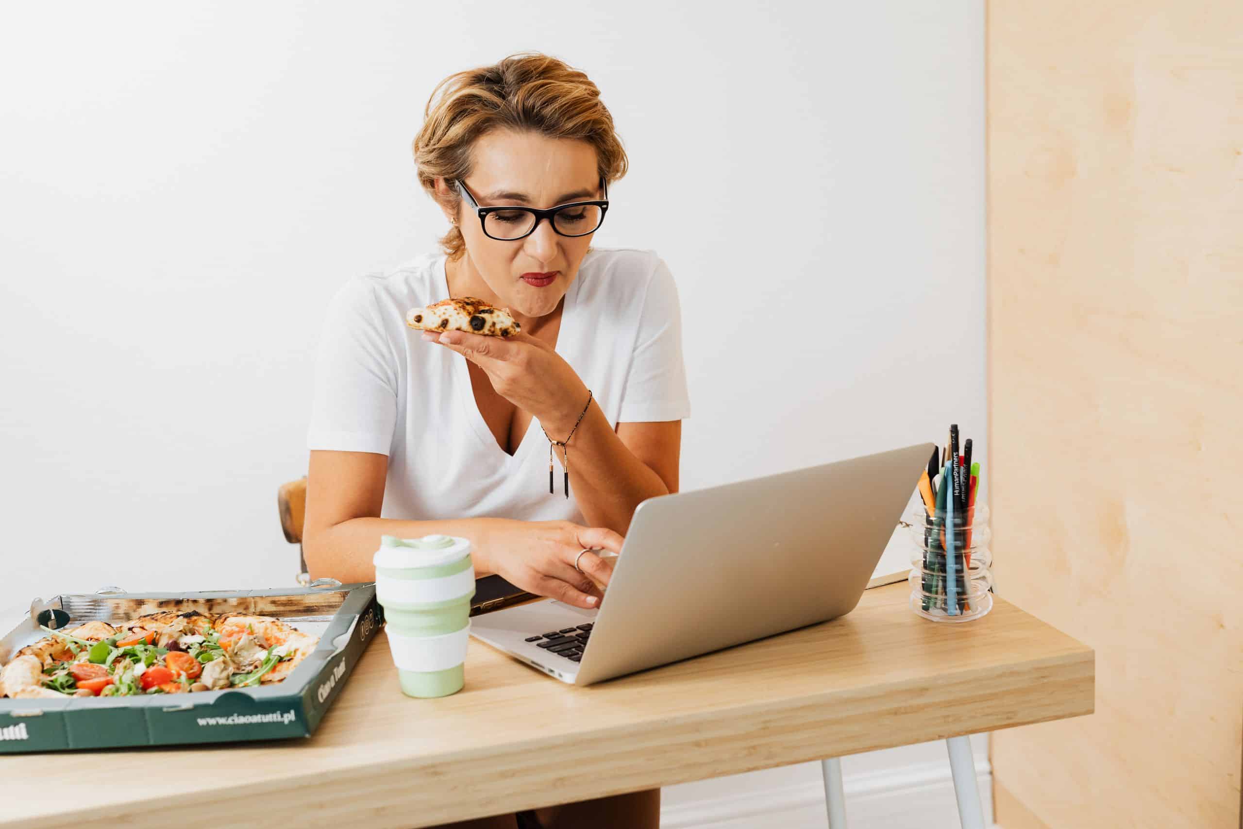 Woman multitasking by enjoying pizza while working on a laptop at home.