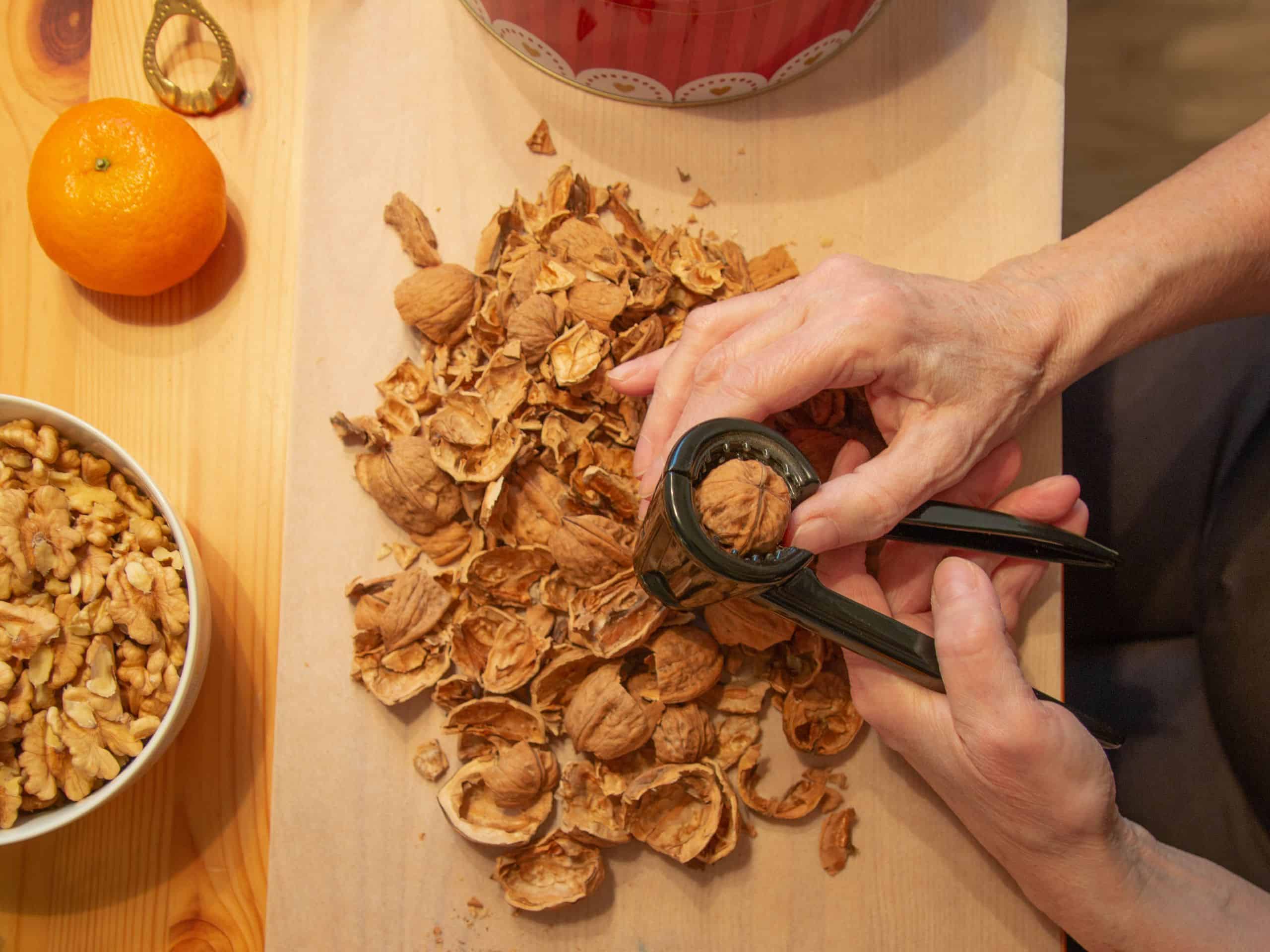 Close-up of hands cracking walnuts with a bowl of shelled nuts and an orange on a wooden table.